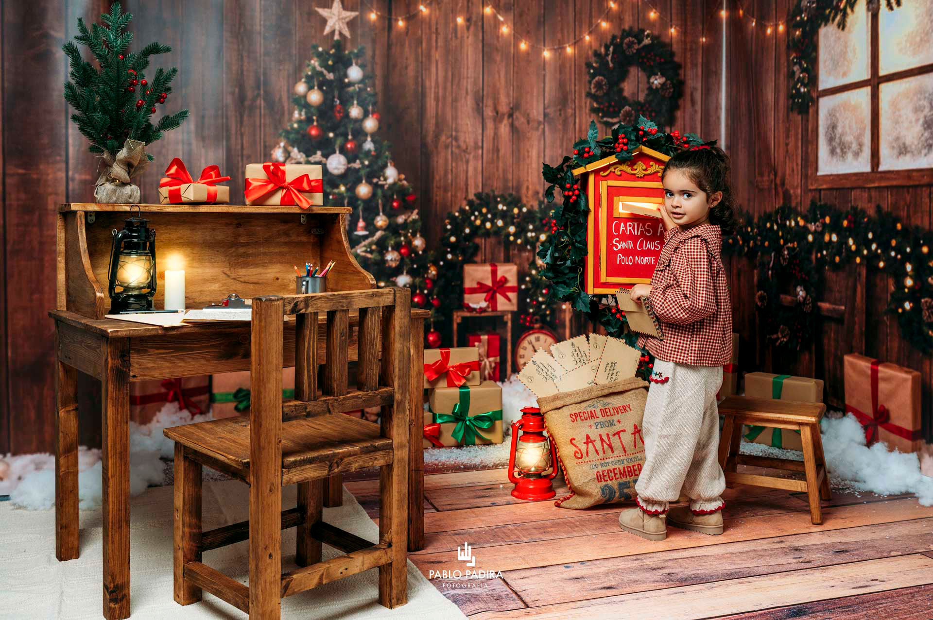 Niña junto al buzón de Papá Noel en un decorado navideño de madera con regalos y luces cálidas.